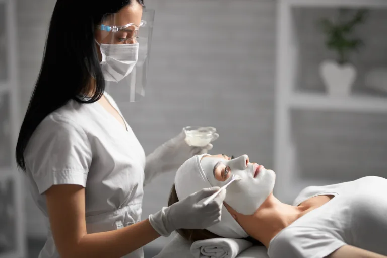 Side view of young beautician in protective mask and rubber gloves doing cleaning face with special cosmetics for young patient. Concept of procedure for improvements and rejuvenation skin.